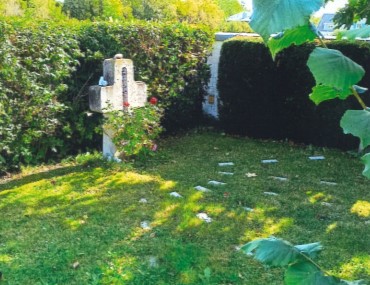 Ein gepflegter kleiner Gartenbereich ist von hohen Hecken umgeben; in einer Ecke steht ein steinernes Denkmal mit Blumen und einer nicht lesbaren Inschrift. Mehrere flache Steine sind auf dem Rasen angeordnet, möglicherweise als Gedenk- oder Grabmarkierungen. Die Szene wirkt ruhig und schattig mit Sonnenlicht, das durch die Bäume fällt.