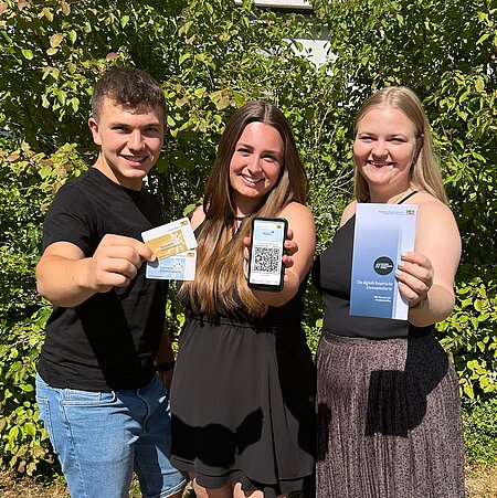 Drei junge Personen halten blaue und goldene Ehrenamtskarte, ein Smartphone und einen Flyer zur Ehrenamtskarte in die Kamera