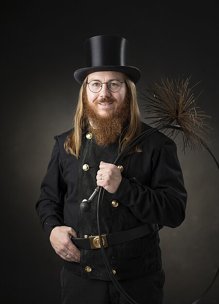 Person in traditioneller Schornsteinfegeruniform mit Zylinder und Rußbesen vor dunklem Hintergrund – ikonische Darstellung eines historischen Berufs mit Glückssymbolik.