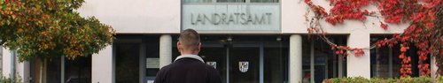 Ein Gebäude mit der Aufschrift „LANDRATSAMT“ über dem Eingang ist zu sehen; davor steht eine Person mit dem Rücken zur Kamera, umgeben von herbstlich gefärbten Pflanzen und Bäumen mit roten und grünen Blättern.