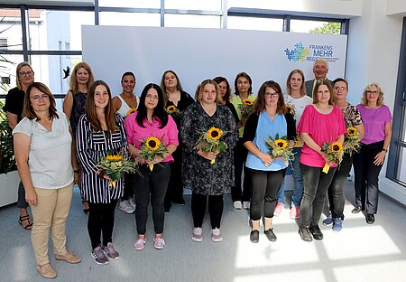 Gruppe von Personen steht in einem hellen Raum vor einer weißen Wand mit dem Logo „Frankens MEHR Region“, mehrere Personen halten Sträuße mit Sonnenblumen in den Händen; im Hintergrund sind große Fenster und Pflanzen sichtbar.