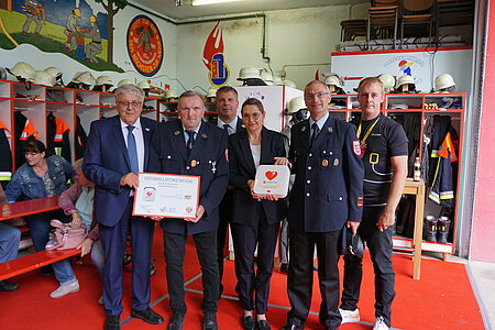Sechs Personen stehen in einer Feuerwache. Vier tragen Feuerwehruniform, eine Person einen Anzug, eine ein schwarzes T-Shirt mit Feuerwehrmotiv. Zwei halten Gegenstände: eine Urkunde mit „DEFIBRILLATORSPENDE“ und eine weiße Box mit rotem Herzsymbol. Im Hintergrund hängen Helme und Uniformen, Wandmalereien zeigen Feuerwehrsymbole.