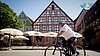 Ein sonniger Moment auf dem Marktplatz: Ein Radfahrer durchquert das Kopfsteinpflaster vor farbenfrohen Fachwerkhäusern, während Gäste unter weißen Schirmen das Flair genießen – Alltag und Lebensfreude in Franken vereint.