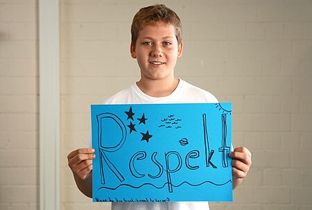 Ein Junge in weißem T-Shirt hält ein blaues Poster mit dem Wort „Respekt“ in großen, dekorativen Buchstaben. Das Poster zeigt Sterne, Wellen, Sonne und Fischmotive. Der Hintergrund besteht aus einer hellen Ziegelwand.