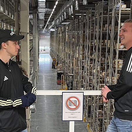 Zwei Personen mit schwarzen Jacken stehen auf einer Plattform über einem großen Lager mit Regalen voller Kartons – zwischen ihnen ein Schild mit dem Hinweis „Nicht auf die Ablagen klettern! Verletzungsgefahr!“, das auf Sicherheitsvorschriften im Lagerbetrieb aufmerksam macht.