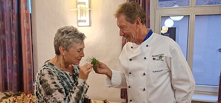 Zwei Personen stehen in einem Innenraum. Eine trägt eine weiße Kochjacke mit blauer Paspel und der Aufschrift „Medizinische Küche – G. Vollkommer“ und reicht der anderen, die ein gemustertes Oberteil trägt, frische Kräuter zum Riechen. Im Hintergrund sind ein gedeckter Tisch, violette Vorhänge und Fenster mit Lichtreflexionen sichtbar.