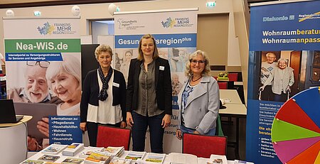 Messestand mit vier Informationsbannern und drei Frauen hinter einem Tisch mit Broschüren – Themen sind Seniorenversorgung, Wohnraumberatung und Gesundheitsregionplus im Rahmen regionaler Sozial- und Gesundheitsangebote.