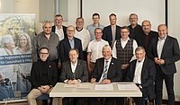 Gruppenfoto der Beteiligten, vier davon sitzen am Tisch und unterschreiben