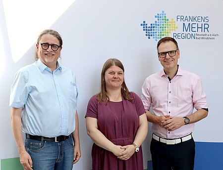Eine Frau und zwei Männer stehen vor einer Fotowand