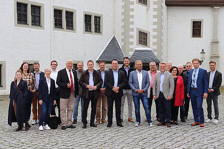 Gruppe von Personen steht auf einem gepflasterten Platz vor einem historischen Gebäude mit weißer Fassade, grauen Schieferdächern und dekorativen Steinrahmen um Türen und Fenster; im Hintergrund sind eine Laterne und architektonische Details sichtbar.
