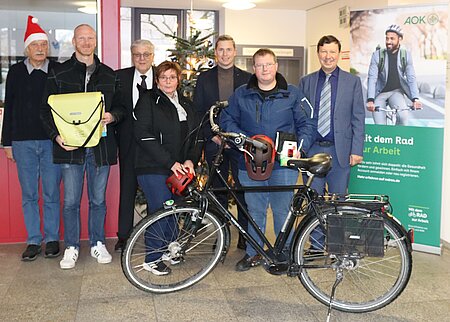 Acht Personen stehen in einem festlich dekorierten Innenraum neben einem Weihnachtsbaum und einem Banner der AOK – eine Person trägt eine Nikolausmütze, eine andere hält eine gelbe Tasche, im Vordergrund ein ausgestattetes Fahrrad. Das Banner wirbt für die Aktion „Mit dem Rad zur Arbeit“ und betont Gesundheit, Umwelt und Kostenersparnis.
