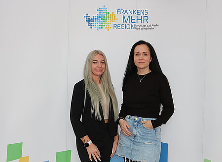 Zwei Personen stehen vor einer weißen Wand, eine trägt einen schwarzen Blazer über einem hellen Oberteil, die andere ein schwarzes Oberteil mit einer hellblauen Jeansrock-Kombination