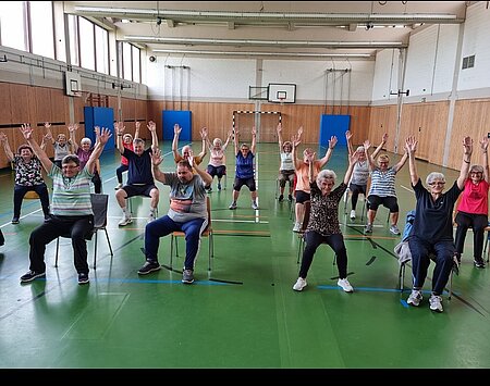 Mehrere ältere Personen sitzen auf Stühlen in einer Turnhalle und führen gemeinsam Armübungen durch. Der Boden ist grün, im Hintergrund sind Holzverkleidung, Basketballkörbe und ein Tor zu sehen. Die Gruppe wirkt konzentriert und aktiv.
