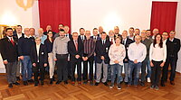 Gruppe von Menschen steht in einem Raum mit Holzfußboden und weißen Wänden, im Hintergrund zwei rote Vorhänge und eine Wandlampe, vorne eine Flasche Wasser auf einem Tisch sichtbar.