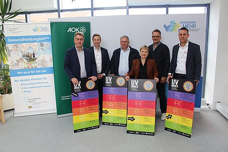 Sechs Personen stehen vor einem weißen Hintergrund mit dem Logo „Frankens MEHR Region“ sowie zwei Roll-Up-Bannern der Gesundheitsregionplus und AOK, und halten jeweils ein großes farbiges Schild mit der Überschrift „UV INDEX“ und einer Skala von 1 bis 11+.