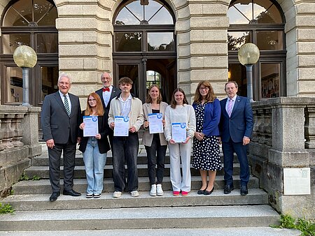 Acht Personen stehen vor einem historischen Gebäude mit Rundbogenfenstern. Vier in der Mitte halten Urkunden. Die Kleidung ist halbformell, das Setting wirkt wie eine Auszeichnungs- oder Abschlussveranstaltung.