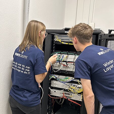 JOBCheckerin Anna-Lena und Azubi Freddy bei HWS Zwei Personen in dunkelblauer Kleidung stehen vor einem offenen Serverschrank und arbeiten gemeinsam an Netzwerktechnik; auf ihren Rücken ist „AWS GRUPPE“ und IT-bezogene Schrift zu sehen.