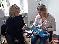 Zwei Personen sitzen in einem Raum auf Stühlen, eine hält eine Babypuppe in blauer Kleidung und gibt ihr mit einer Flasche zu trinken, die andere hält ein kleines Behältnis in der Hand, auf dem Tisch liegen verschiedene Gegenstände.