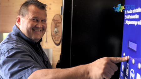 Mann lächelt und bedient blauen Automaten mit deutscher Anleitung: „Produkte auswählen“, „Geld einwerfen“, „Ausgabemenge wählen“ – Szene zeigt Interaktion mit Verkaufs- oder Ticketgerät vor Holzfassade.