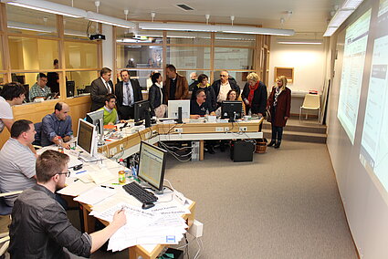 Viele Personen sitzen an einem halbrunden Tisch, einige stehen dahinter und sehen sich eine Präsentation an
