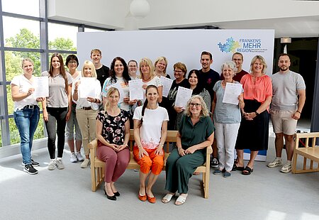 Eine Gruppe von circa 18 Erwachsenen posiert in einem Innenraum vor einem Hintergrund mit der Aufschrift „FRANKENS MEHR REGION“. Einige Personen sitzen in der ersten Reihe, andere stehen dahinter. Mehrere halten Urkunden oder Dokumente in den Händen. Der Raum ist hell und modern, mit großen Fenstern. Die Kleidung reicht von leger bis business-casual.
